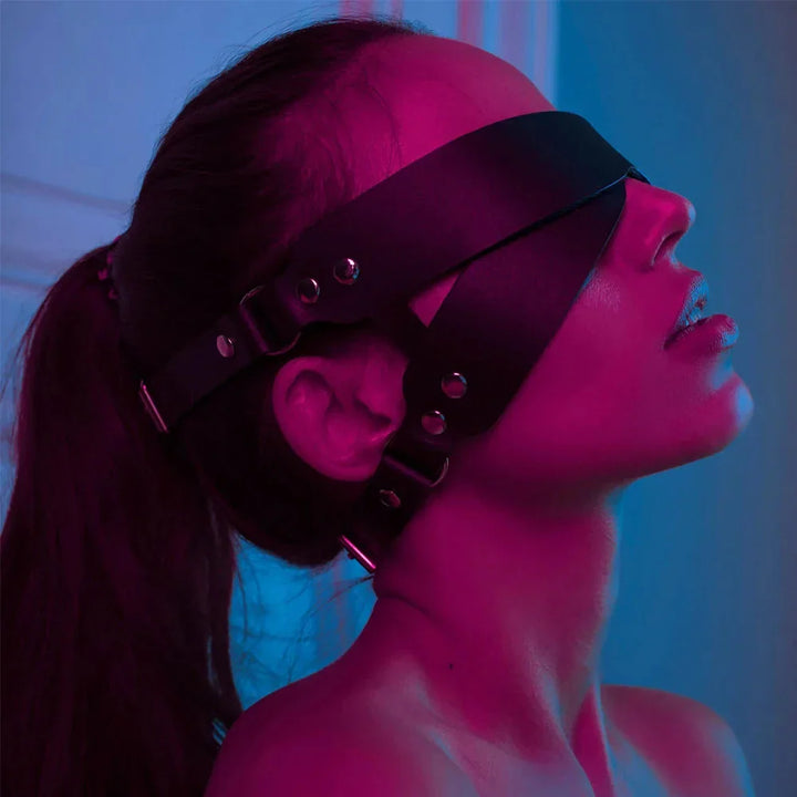 Side view of a woman wearing a black leather blindfold mask with adjustable straps in moody lighting